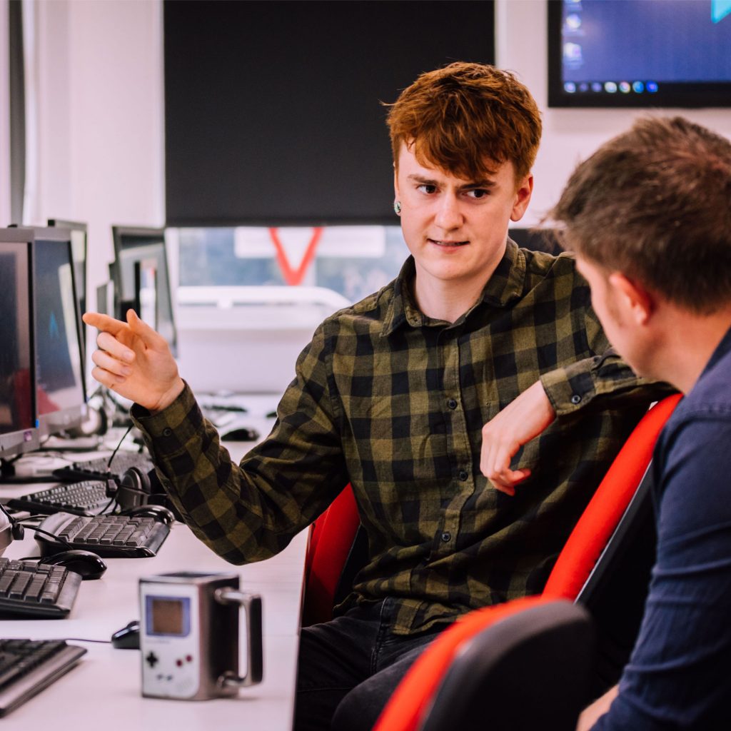 Young man in checked shirt talking to a a colleague in a computer room