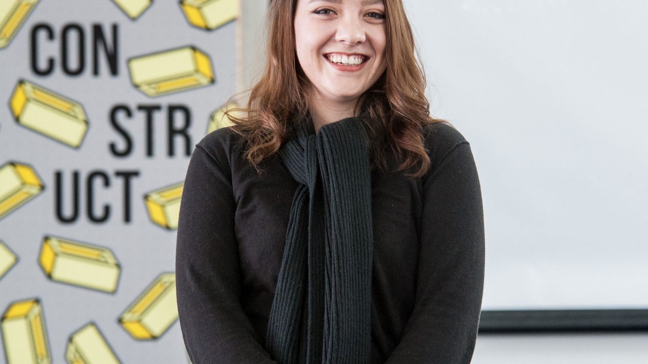 Young woman with brown hair in a black jumper stood next to an advertising poster