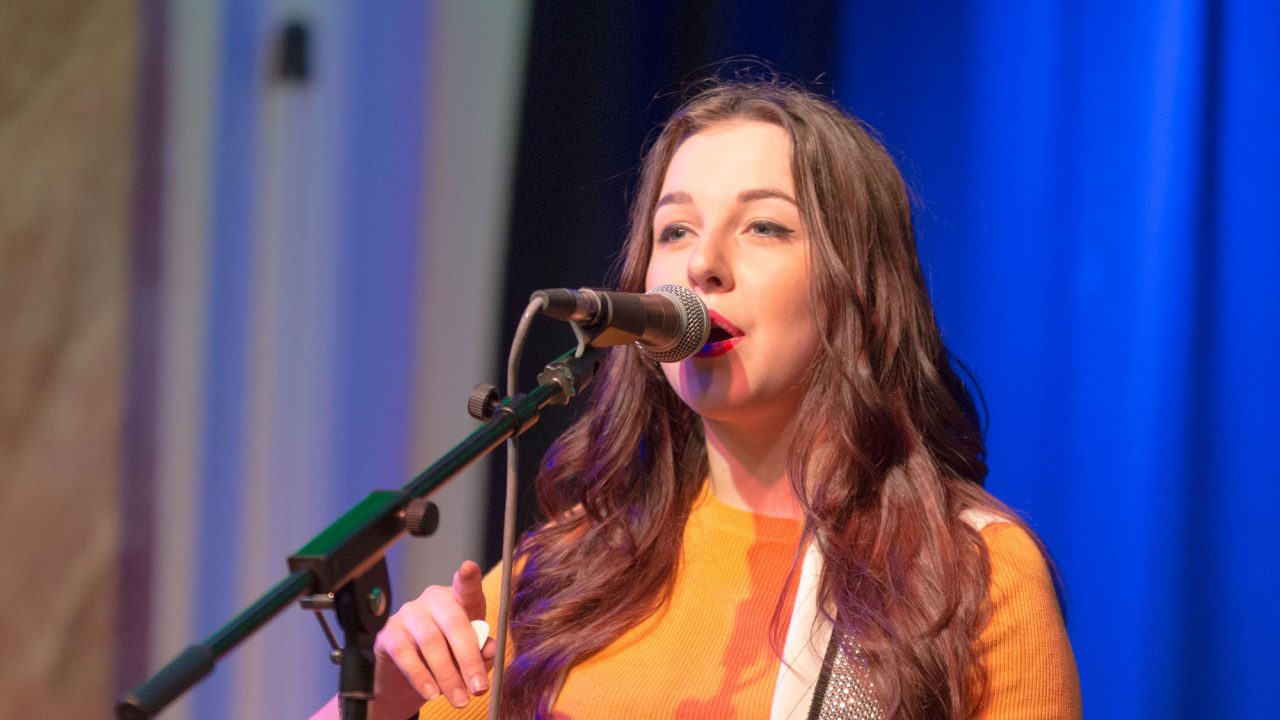 Young woman on stage singing into a microphone and playing an acoustic guitar