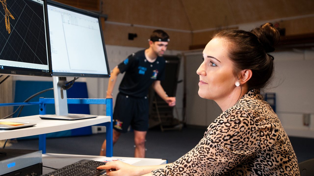 san – sport and exercise – Sports Performance Analysis (MSc) A student on the Sports Performance Analysis course looking at data on a monitor whilst another student wears biometric equipment in the background
