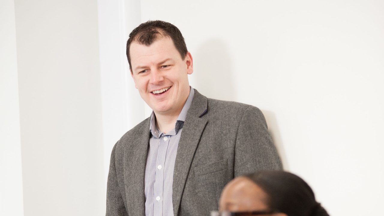 A man holding a cup and smiling whilst a woman is sitting at a laptop and smiling