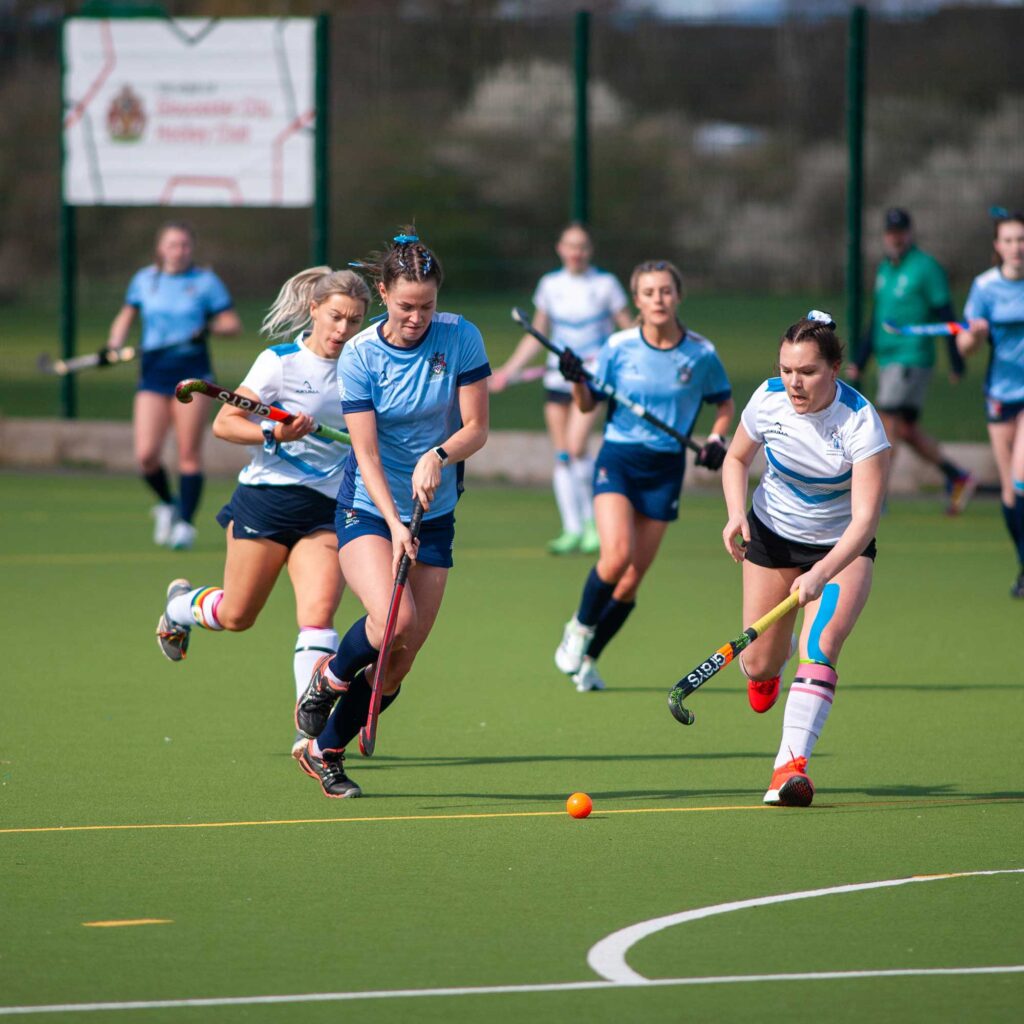 Female hockey players on the AstroTurf chasing after the ball.