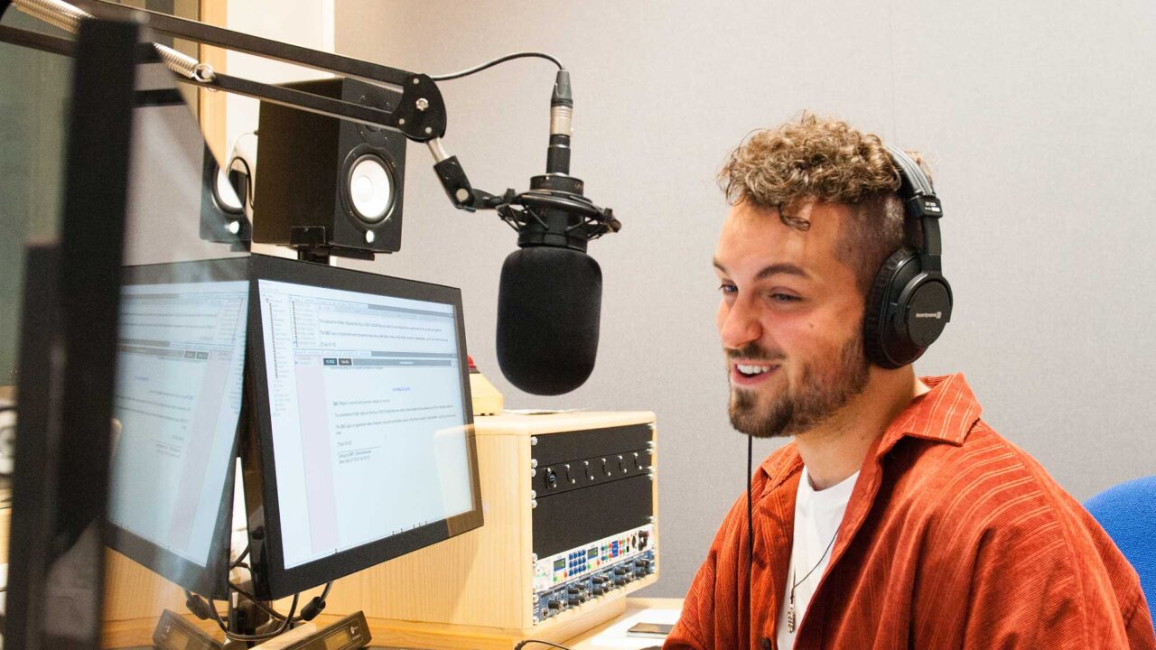 journalism-hero Student sitting in front of mixing desk in a radio studio. He's wearing headphones and talking into a microphone.
