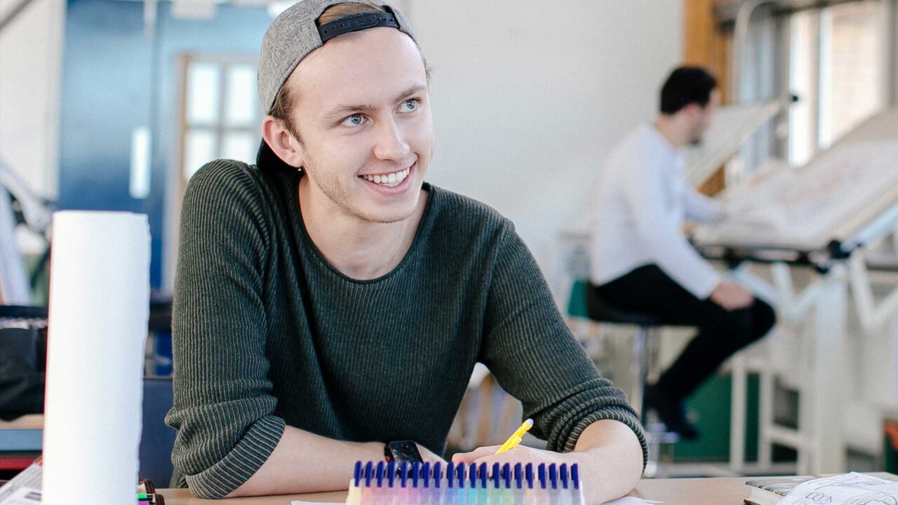 A student sits at a desk with a pencil and illustration work in front of him.