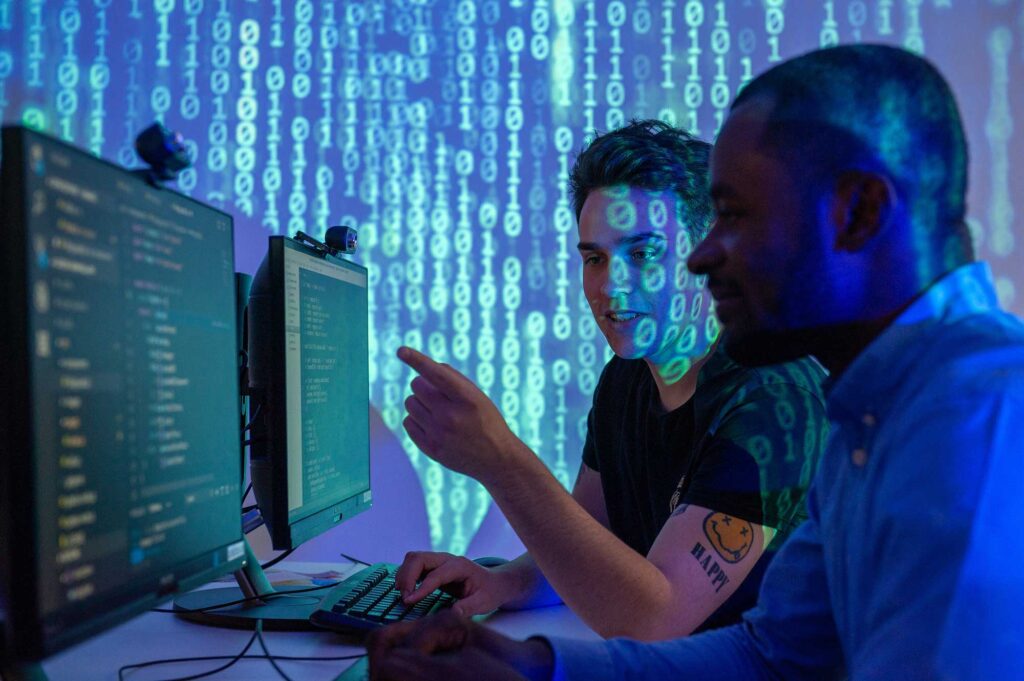 Two students look at computer screens together with lighting showing binary numbers projected onto them.
