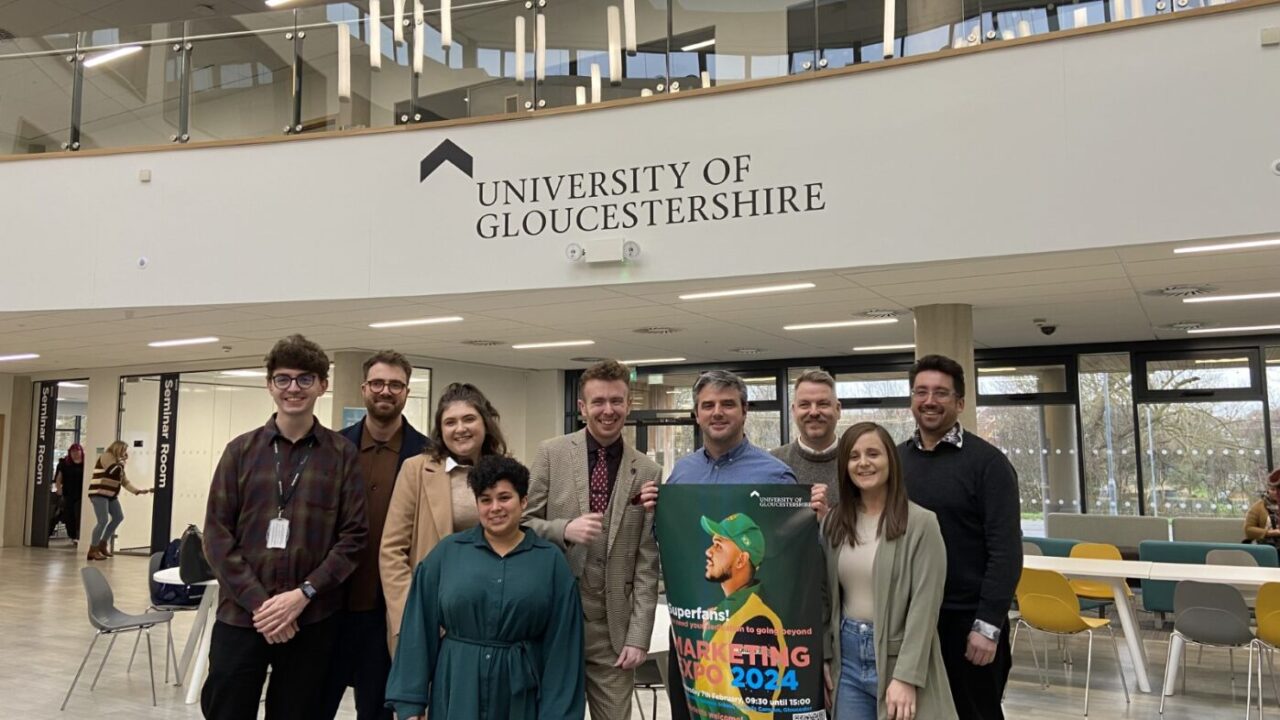 Nine people (staff and alumni) standing in a line and facing the camera smiling with one holding a poster publicising Marketing Expo 2024