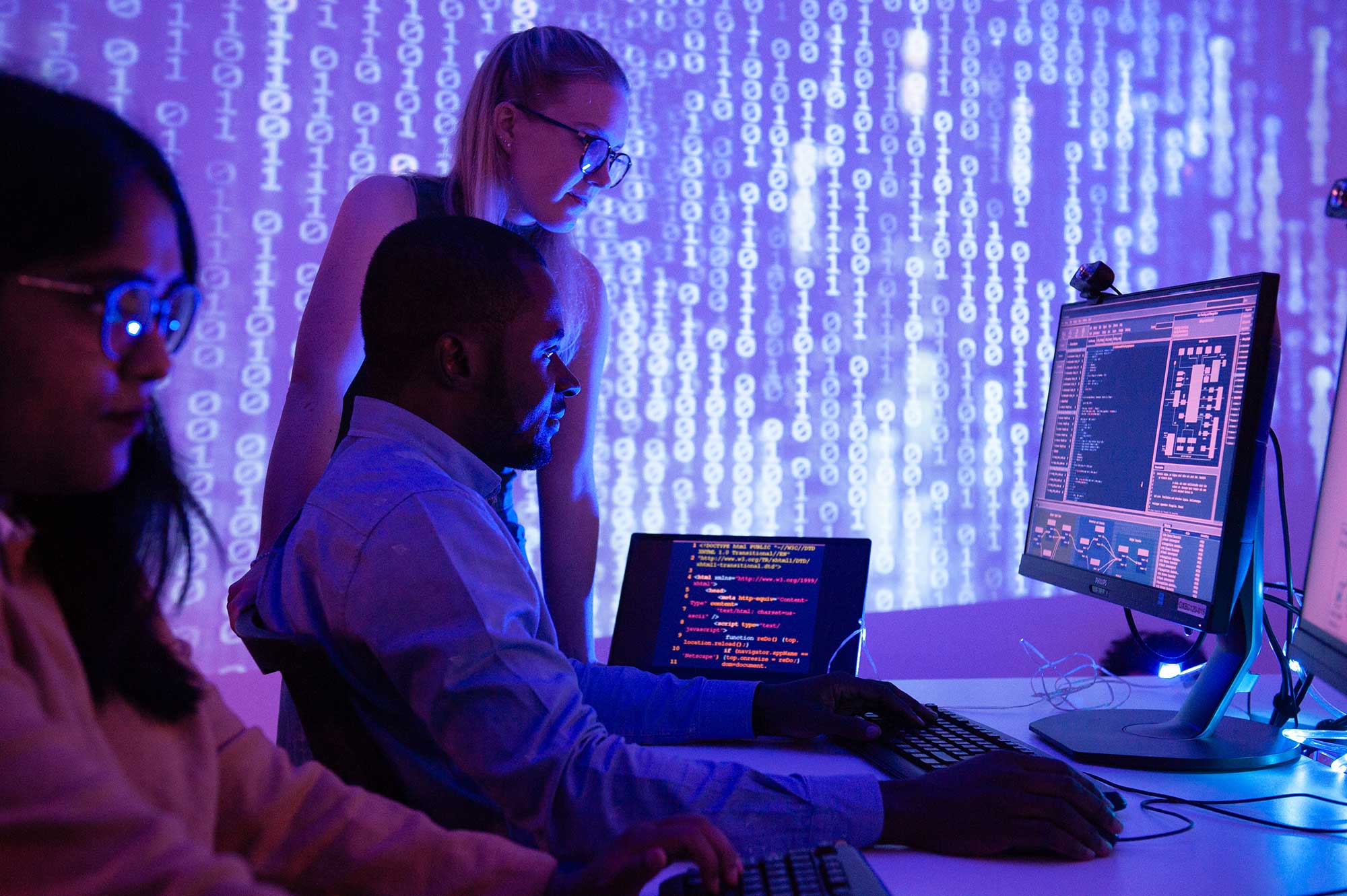 Students sit at computer screens displaying code with a backdrop showing binary numbers.