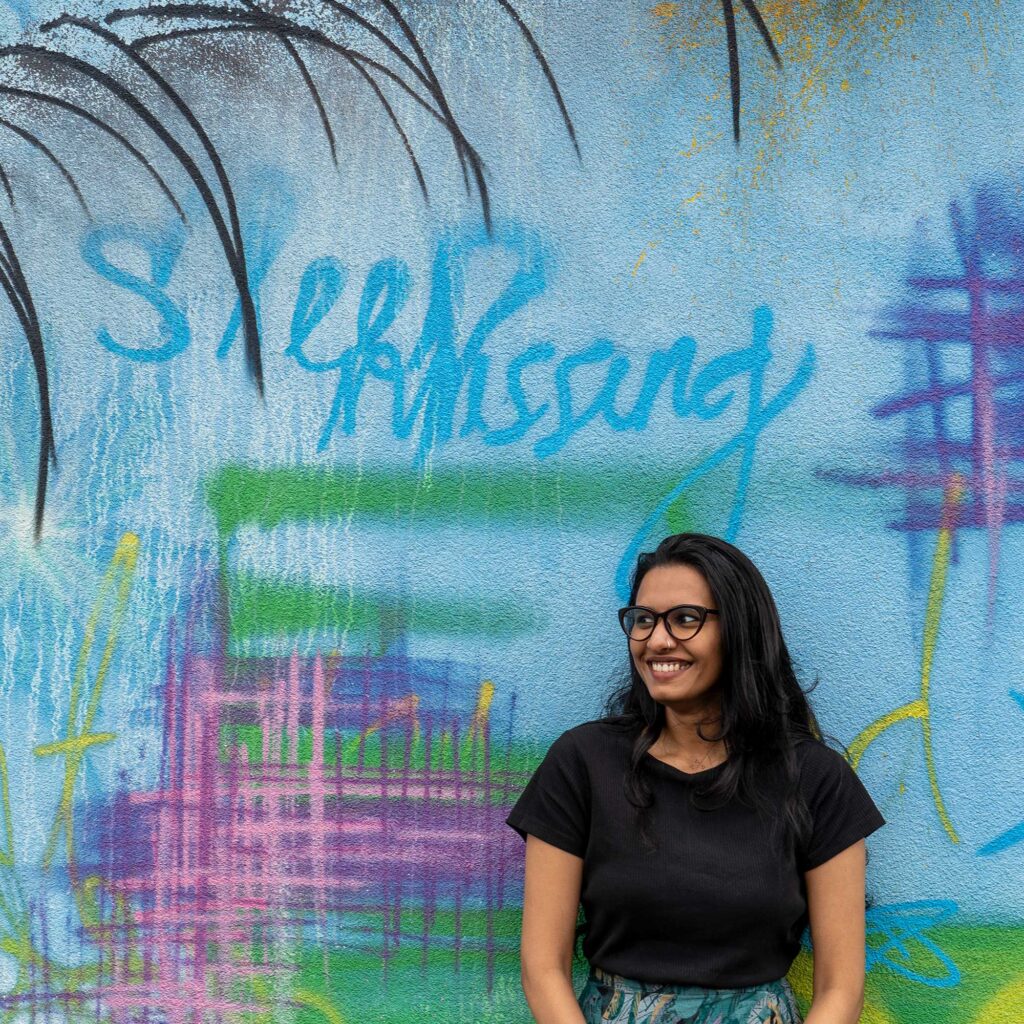 A female student wearing glasses, standing in front of a wall with brightly coloured graffiti on it.