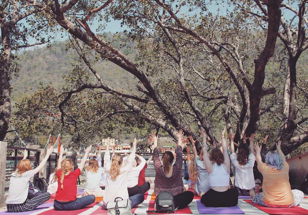 A group of students sit on a mat with arms raised overlooking the Ganges.