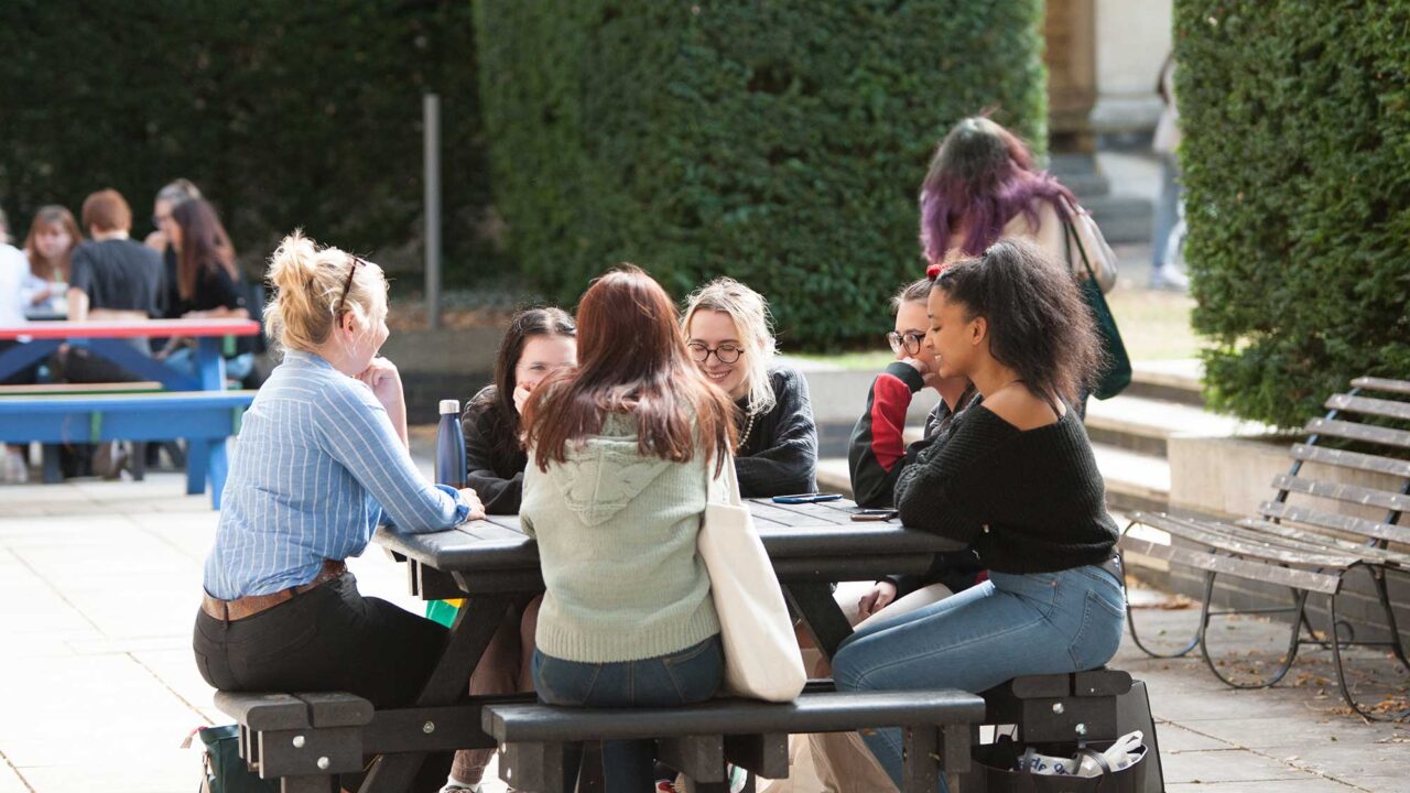 A group of student sit at a wooden picnic table in the grounds of Francis Close Hall.