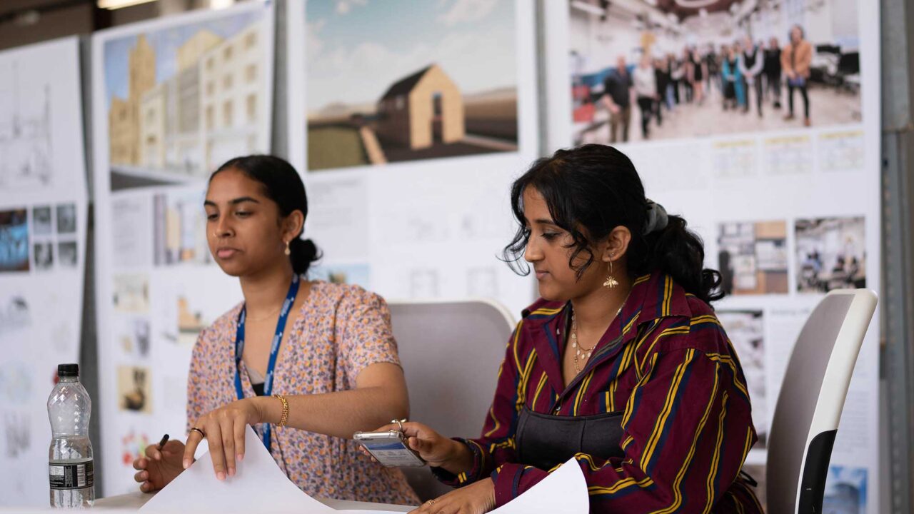 Two students sit at a desk looking at paper plans.