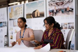 Two students sit at a desk looking at paper plans.