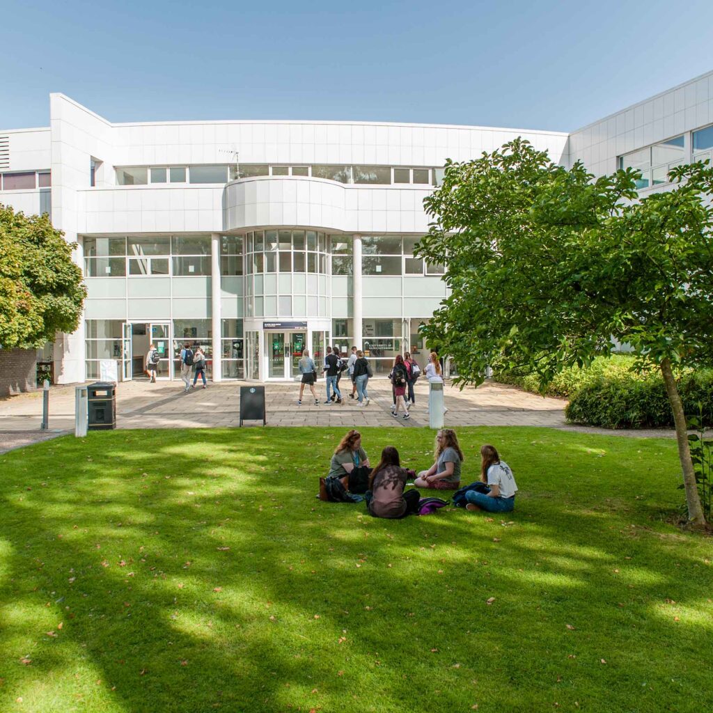 Photo of students sat on the grass at Park Campus, with the main reception in the background.