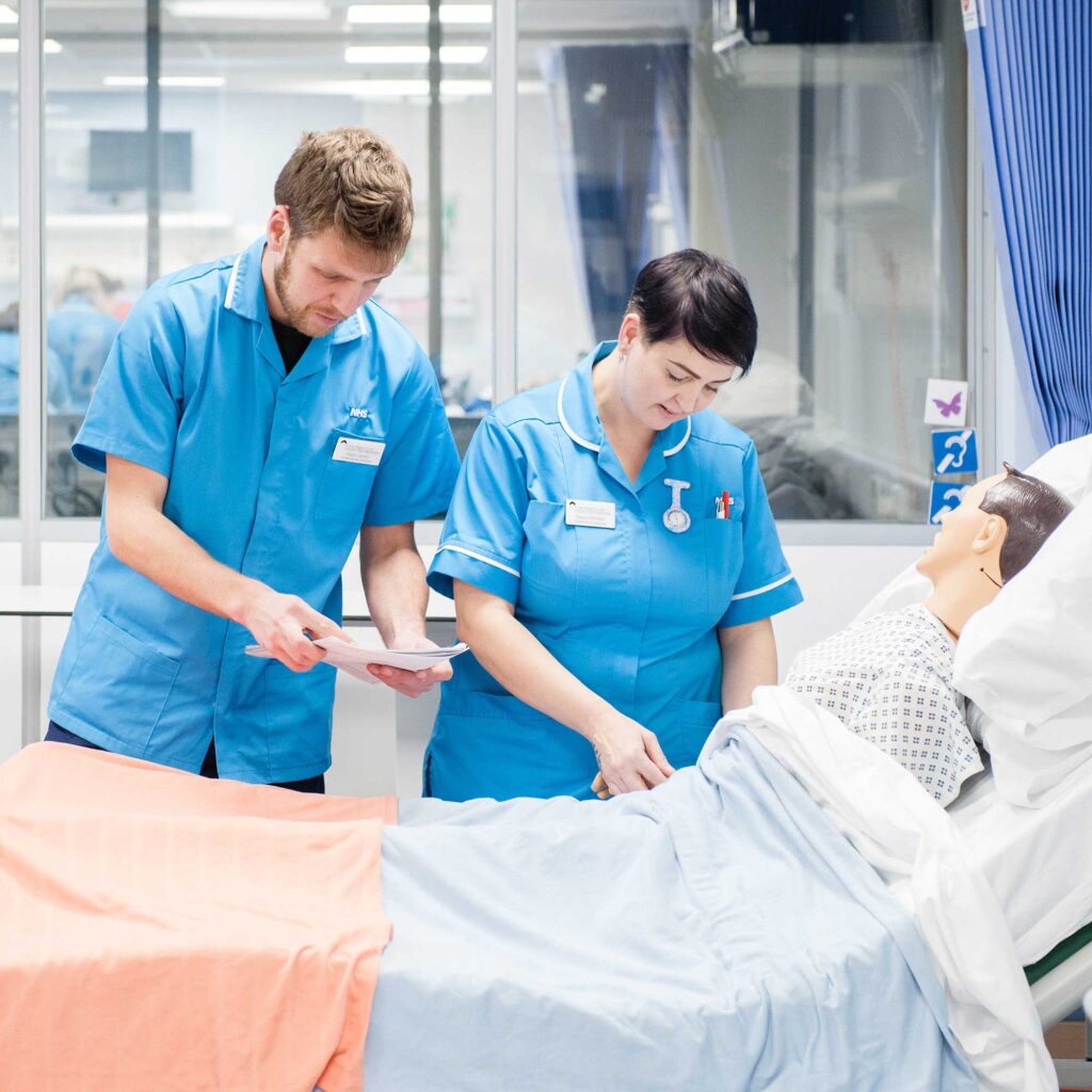 Two Nursing Associate students check the obs of a dummy patient lying in a hospital bed.