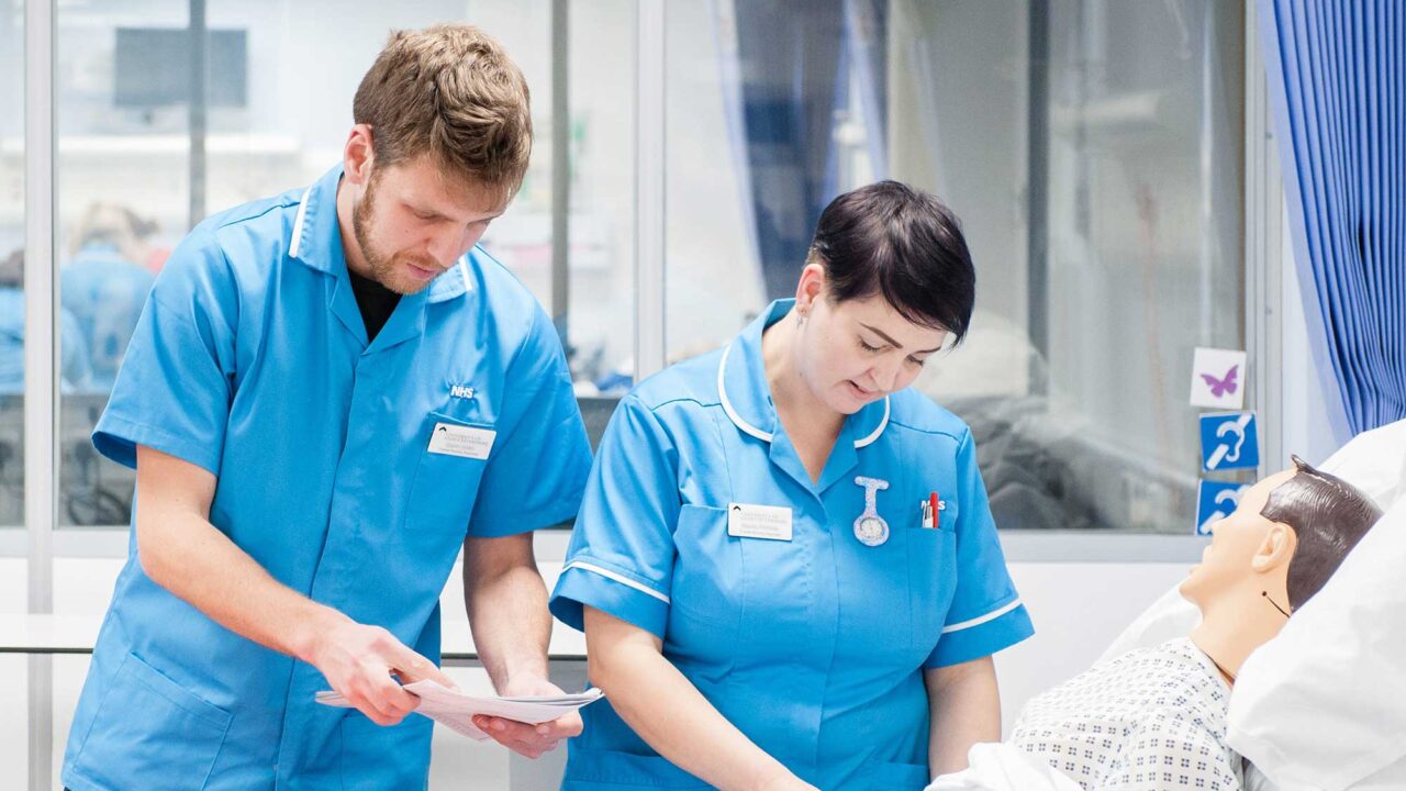 Two Nursing Associate students check the obs of a dummy patient lying in a hospital bed.