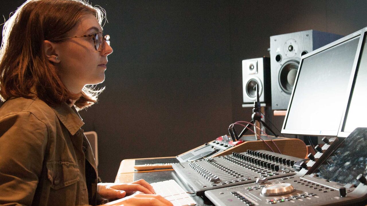 A woman sits at a TV mixing desk with her hand on a mouse.