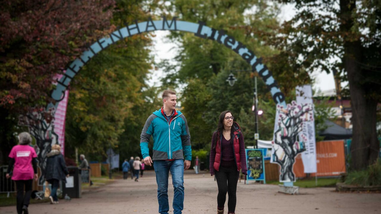 Two students walking in Cheltenham with Cheltenham Literature banner behind them.