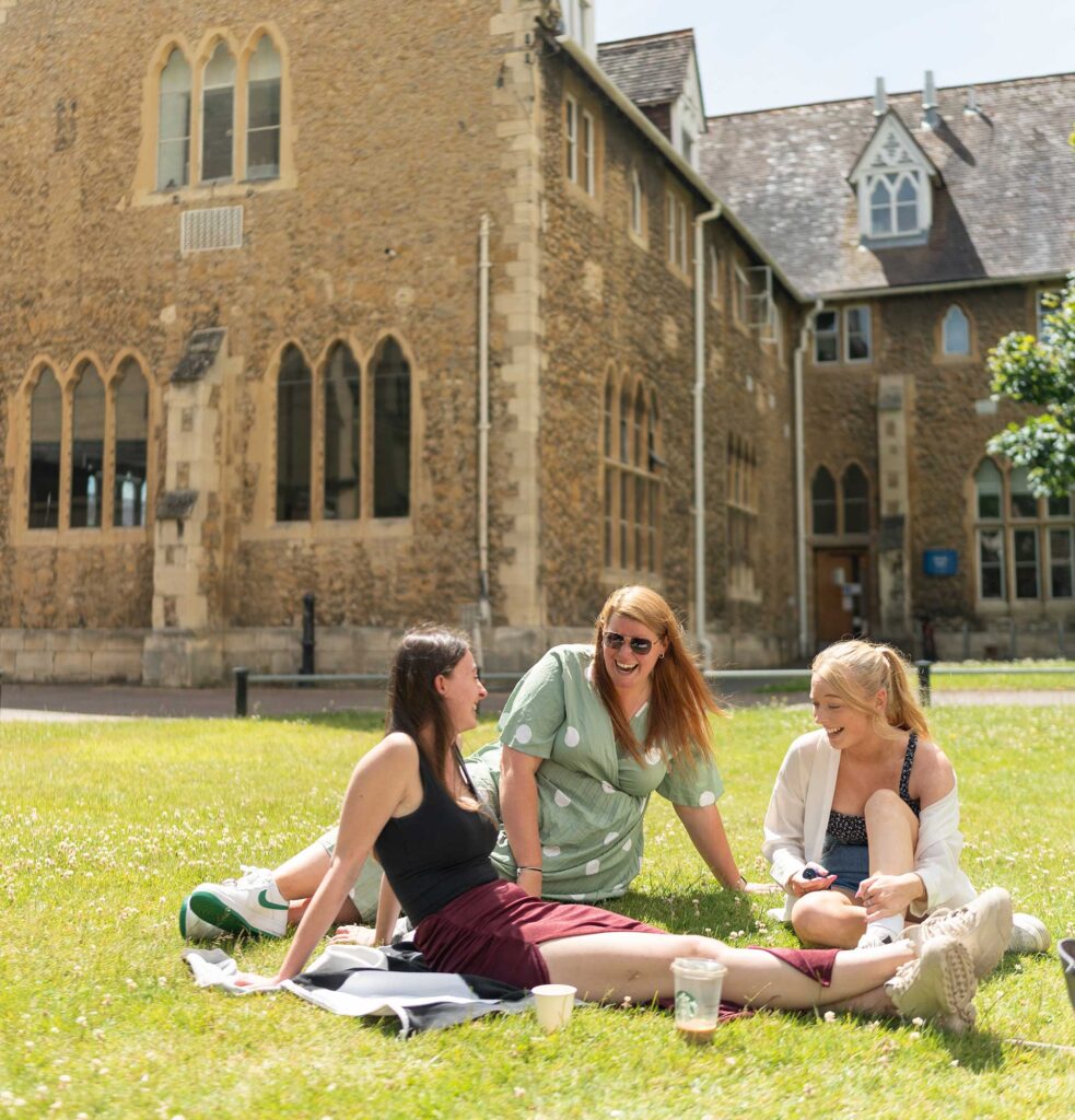 Three students sitting on the Chapel lawn on FCH campus, with the Chapel in the background. The sun is shining and they're smiling and laughing.