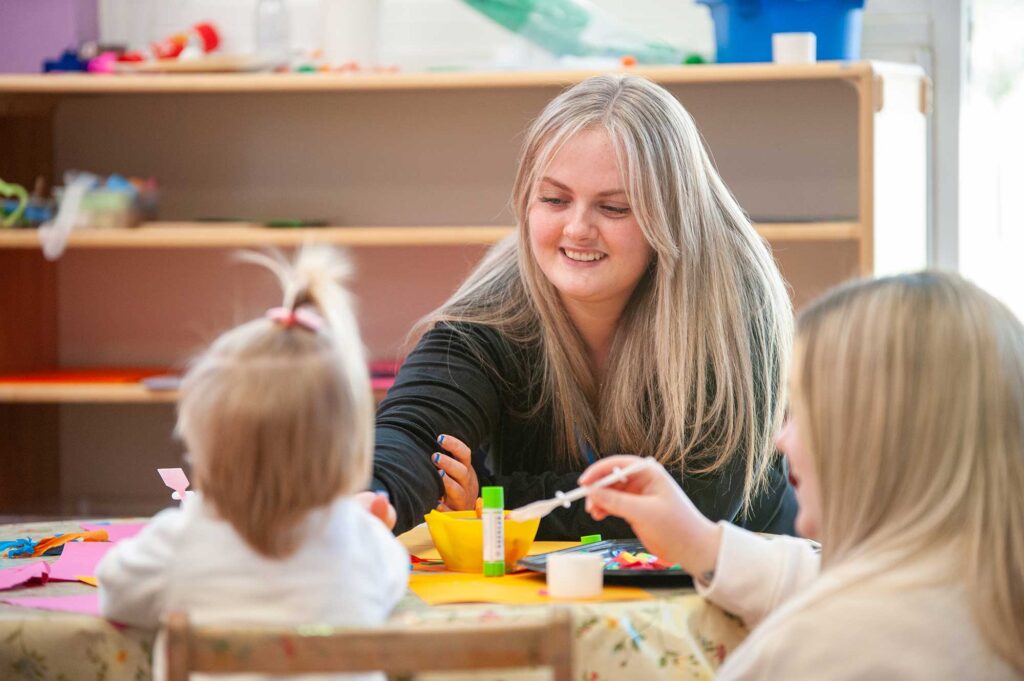A student sits with young children who are involved in a craft activity.