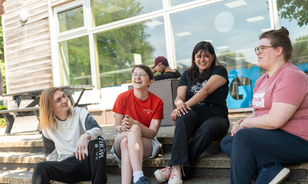 Four students sat on steps outside in sunshine smiling and laughing