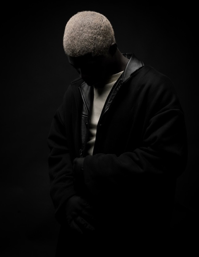 Man looking down at the floor, dressed in black and white, with shadow across his face making bleach blonde hair the focus.