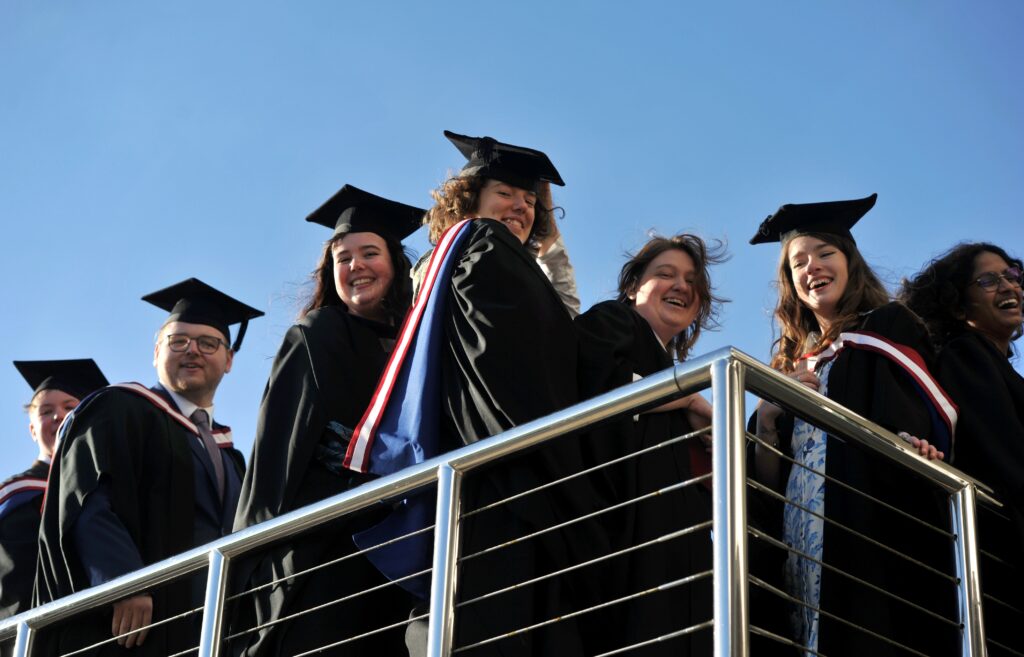 Picture of 7 smiling University of Gloucestershire graduates looking over a balcony at Cheltenham Racecourse on a sunny day. They are wearing graduation hats and gowns.