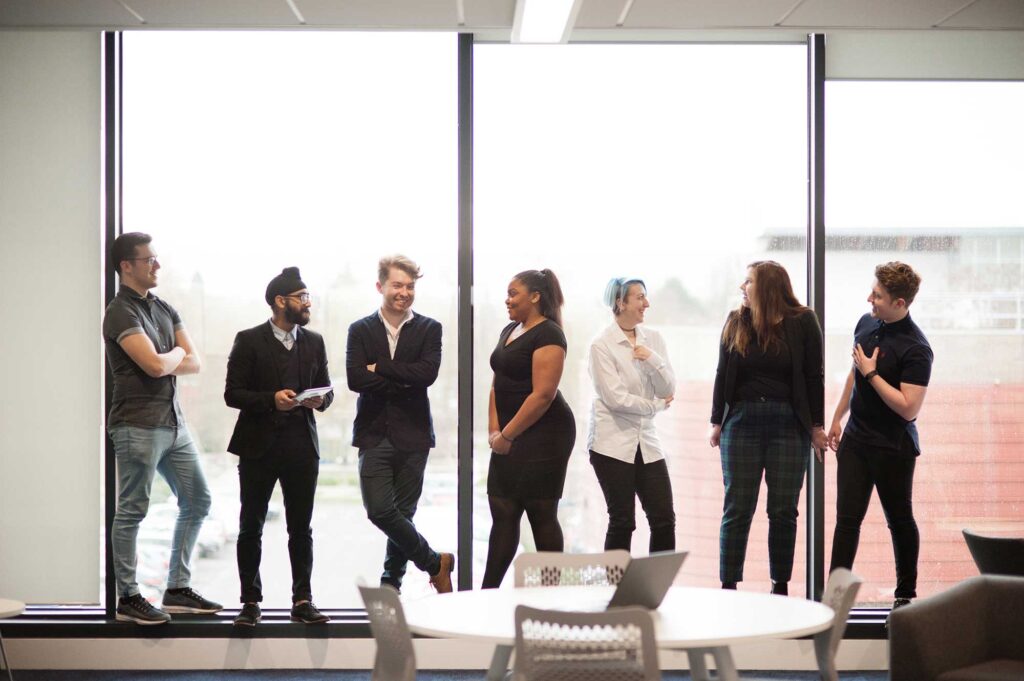 Students standing in a line in front of a window talking.