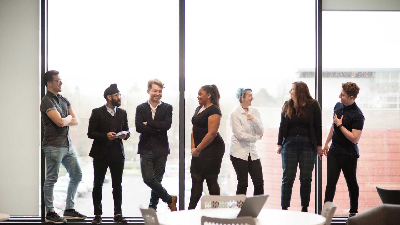 Students standing in a line in front of a window talking.