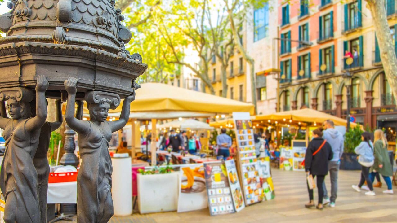 Las Ramblas market in Barcelona, showing a statue in the foreground and people browsing the market in the background.