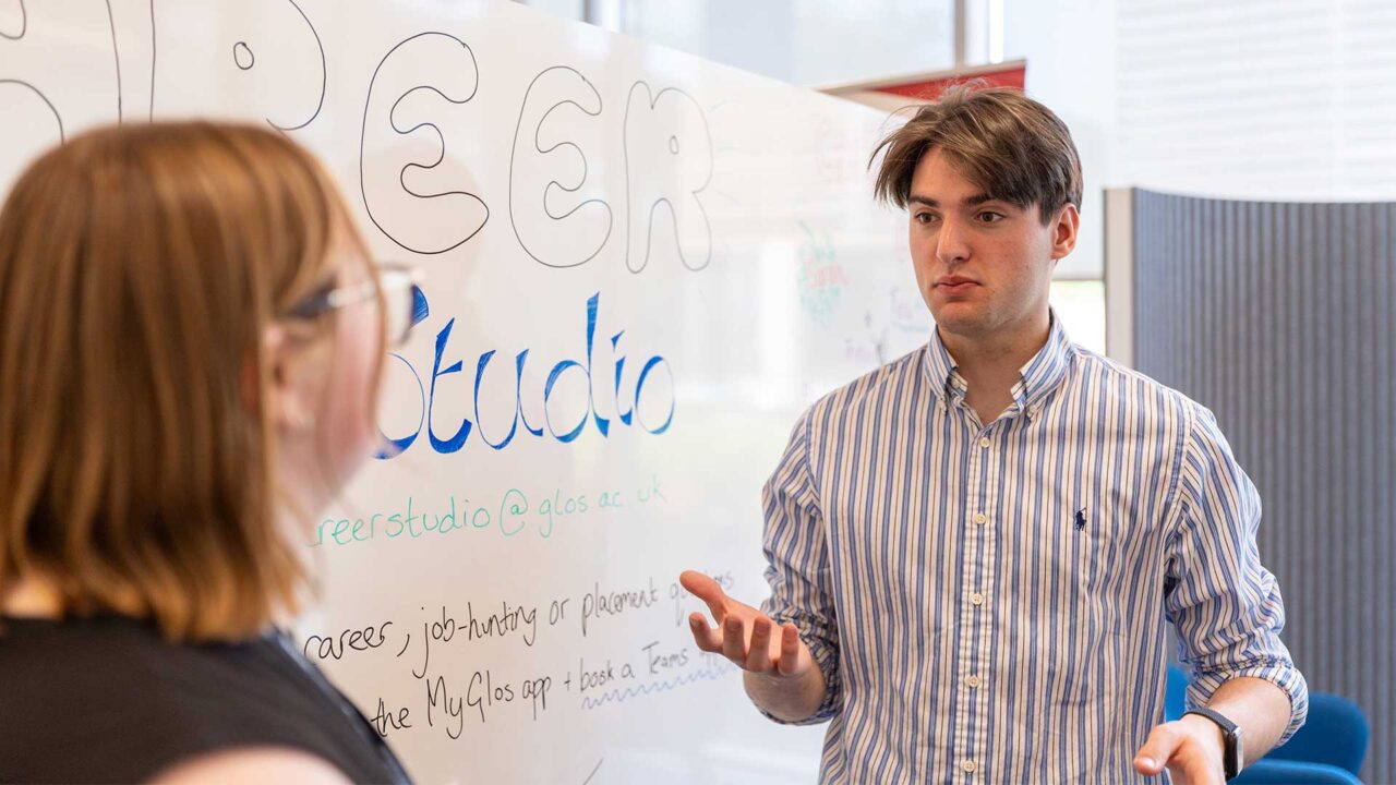 Student and member of staff chatting in front of a board promoting career studio's offer.