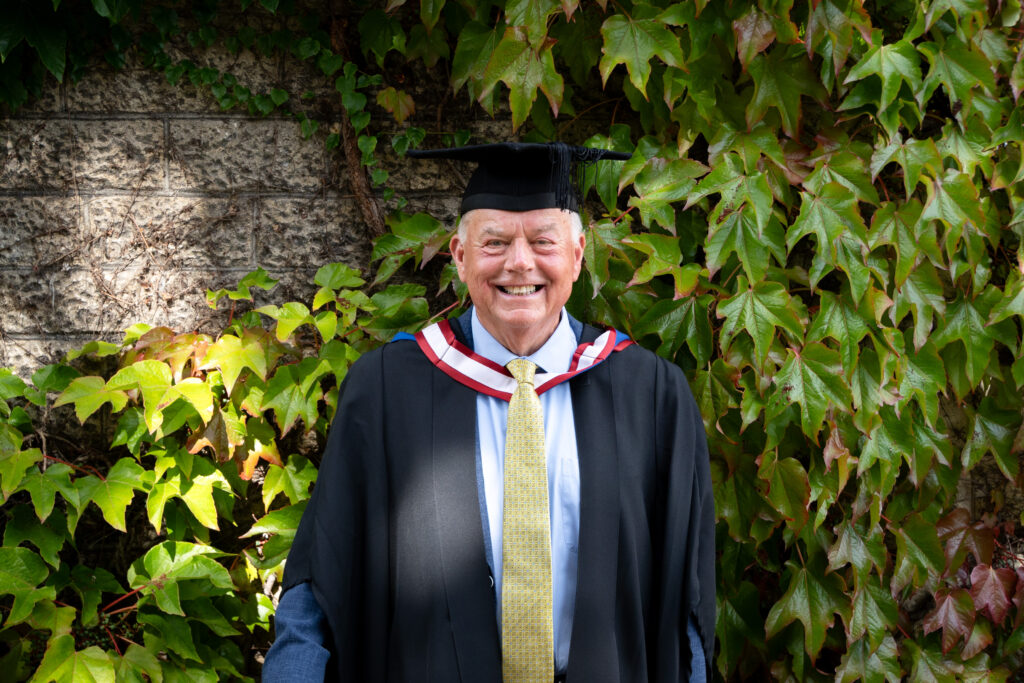 Peter Mothersole dressed in graduate cap and gown outside Francis Close Hall Campus smiling.