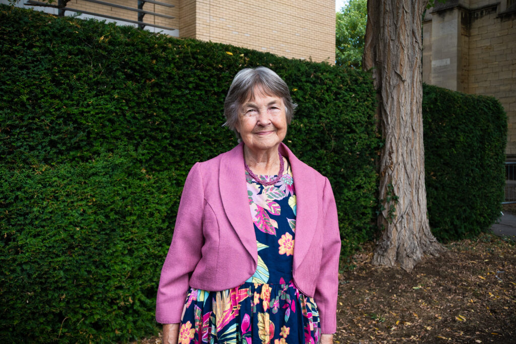 Sandra Walton OBE standing under a tree on the grounds of Francis Close Hall Campus, wearing a navy flower dress and pink jacket.