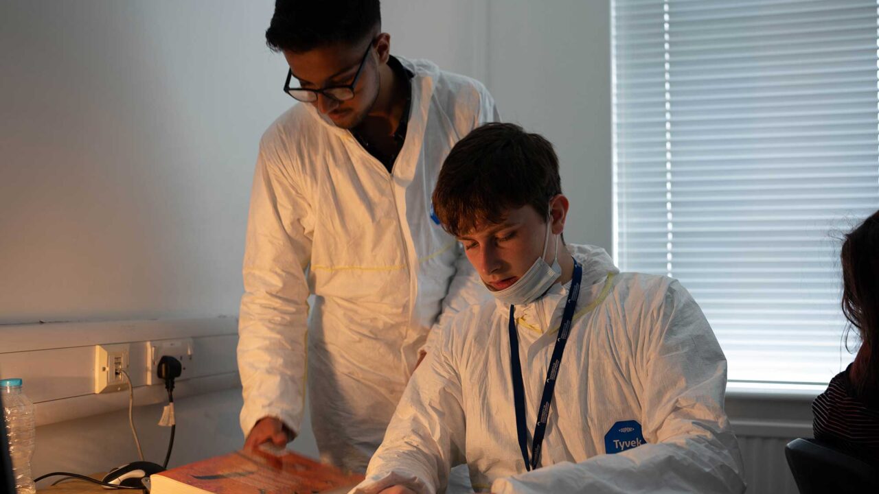 Two students wearing white overalls looking at evidence in an office.