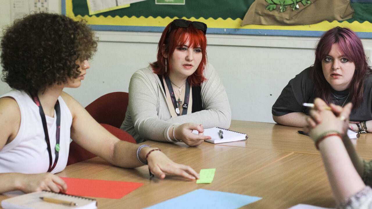 Three students sitting at desks discussing a topic together.