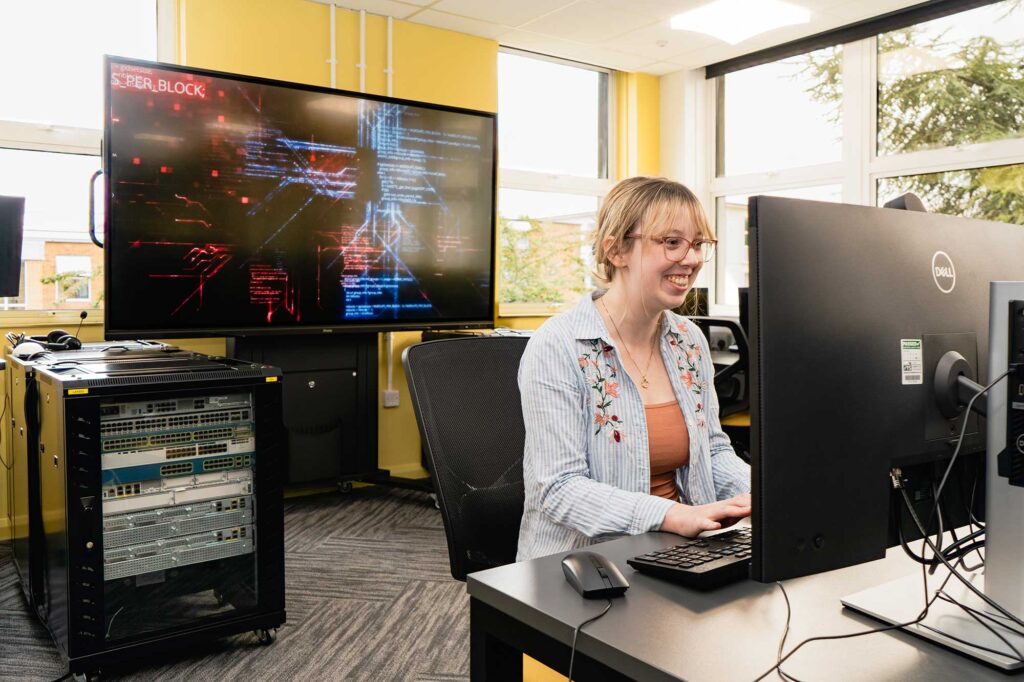 A student using computing equipment in a room with servers and a large digital screen.