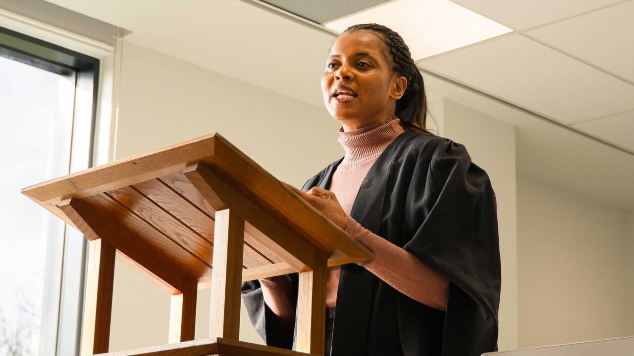 A student standing up at a lectern in a robe, making a case.