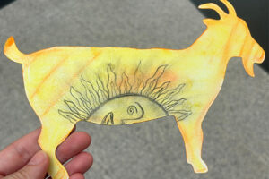 Someone holding a goat postcard decorated in yellow, with orange accents and a half sun face in the belly of the goat.