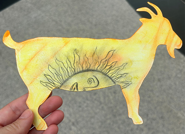 Someone holding a goat postcard decorated in yellow, with orange accents and a half sun face in the belly of the goat.