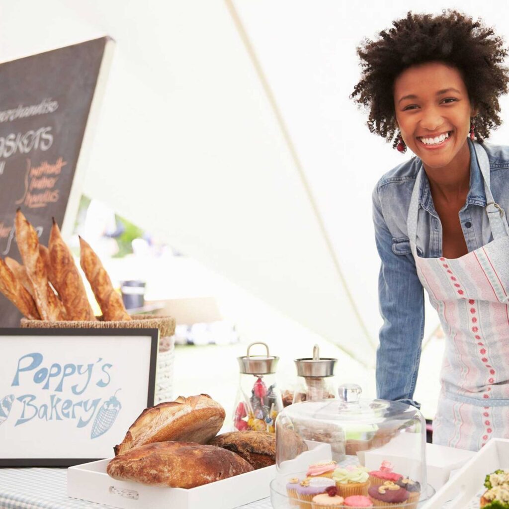 A woman stood at a stand selling baked goods including bread and cakes, with the sign "Poppy's bakery" on the table.
