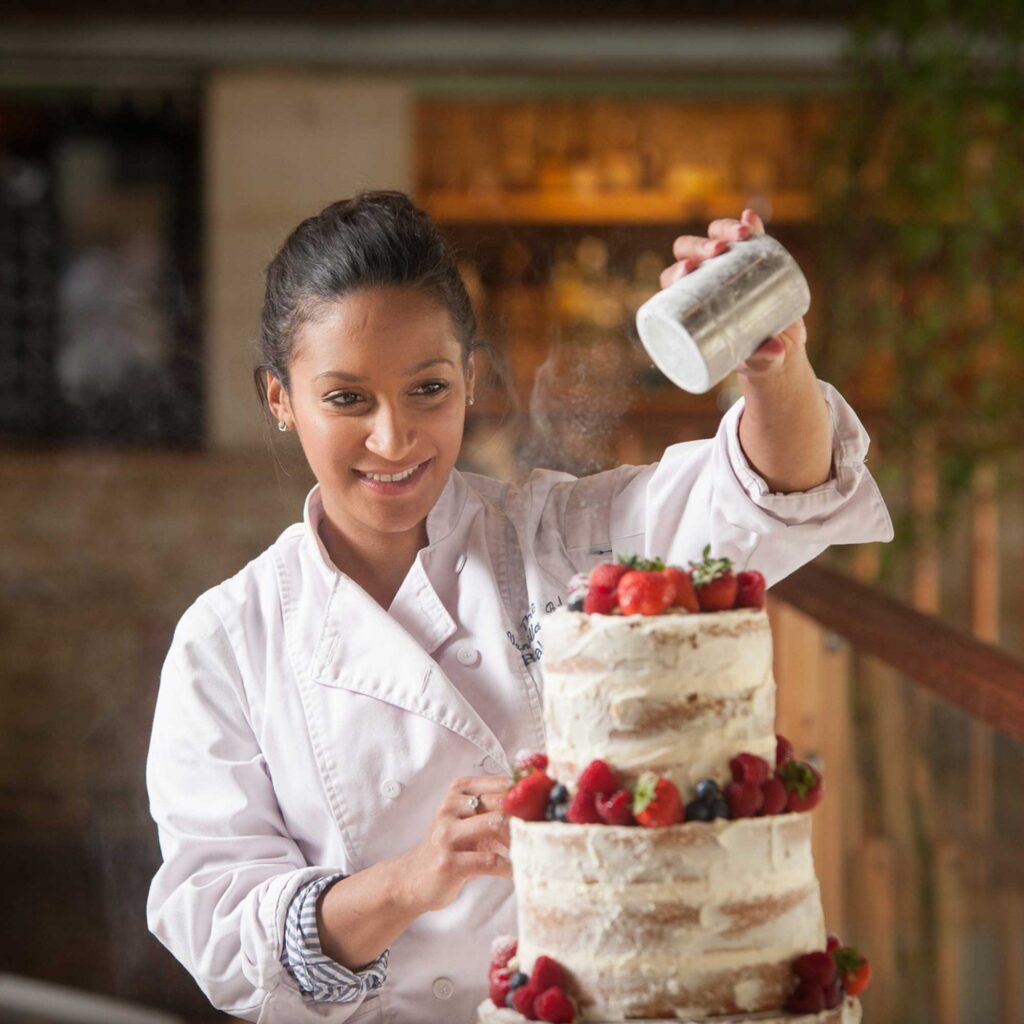 A baker finishing off a fresh fruit cake with an icing sugar shaker.