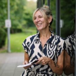 Photo of Hannah Lowe, leaning against a glass door holding a book and smiling.