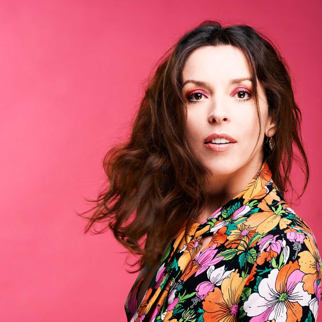 Bridget Christie looking at the camera against a bright pink background.