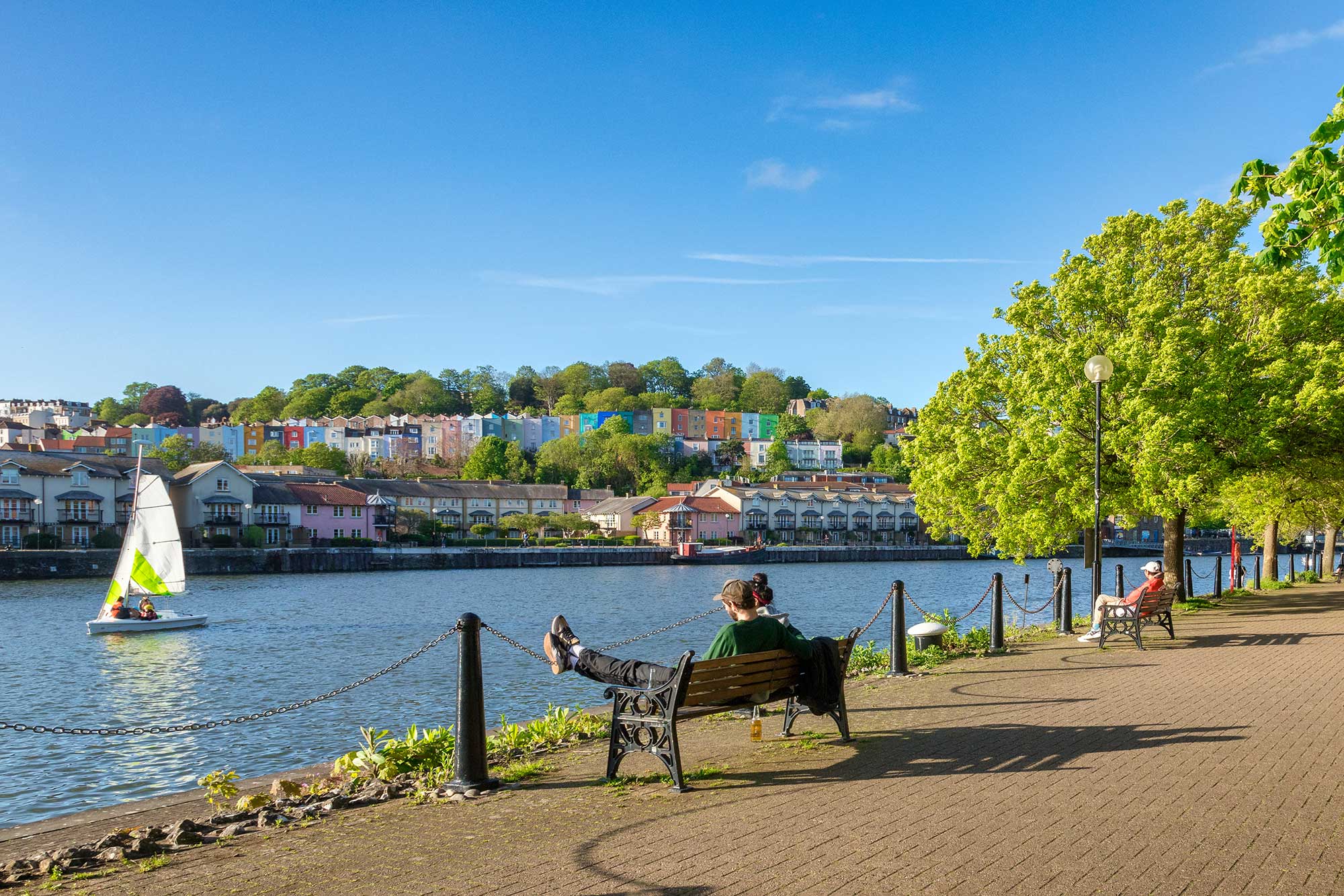A couple sit on a bench overlooking the river in Bristol with colourful houses in the distance.