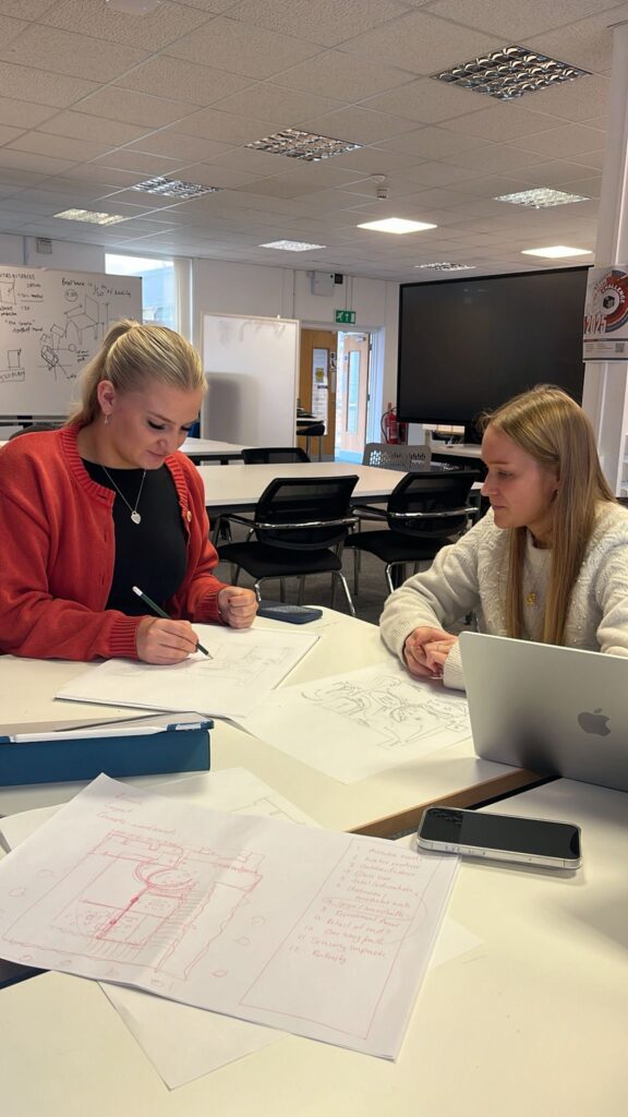 2 students sitting at a table drawing a floor plan, with a sketch in the foreground.