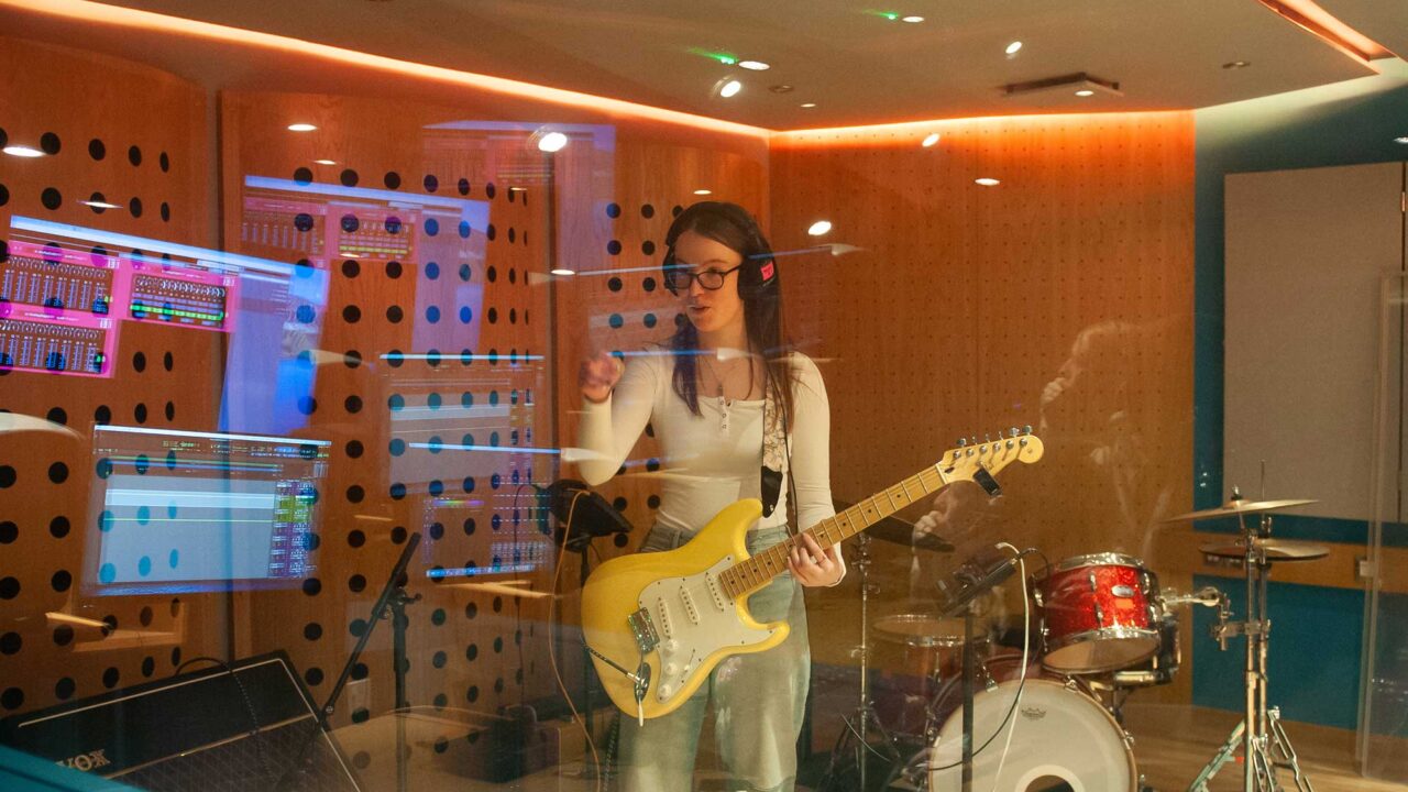 A music student wearing headphomes and hloding a guitar stands behind glass in a recording studio.