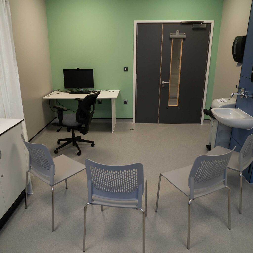 Consulting room at City Campus set up with a desk and computer, 4 chairs and a sink in the room.
