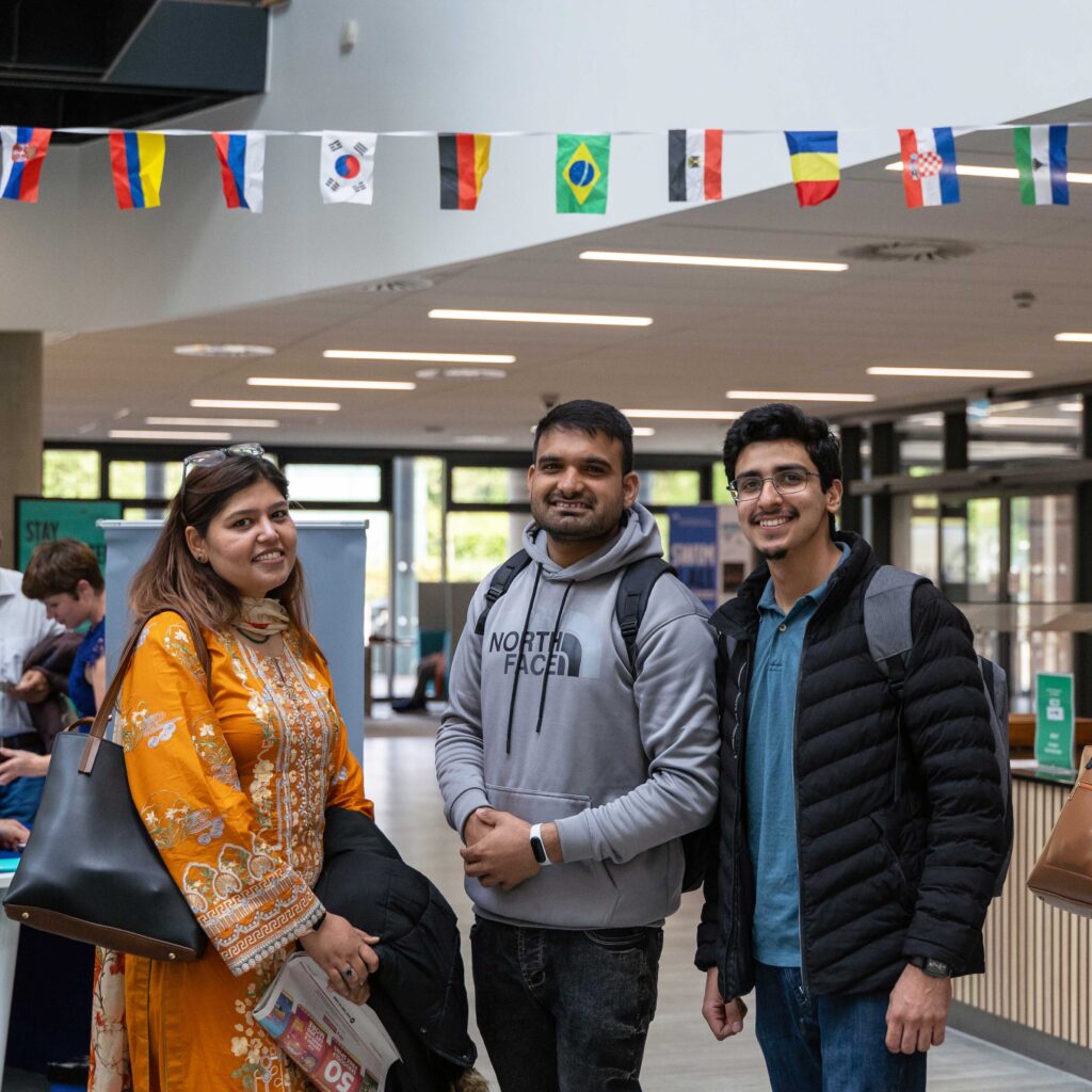 Three international scholars standing in the Business School atrium with flags above them.