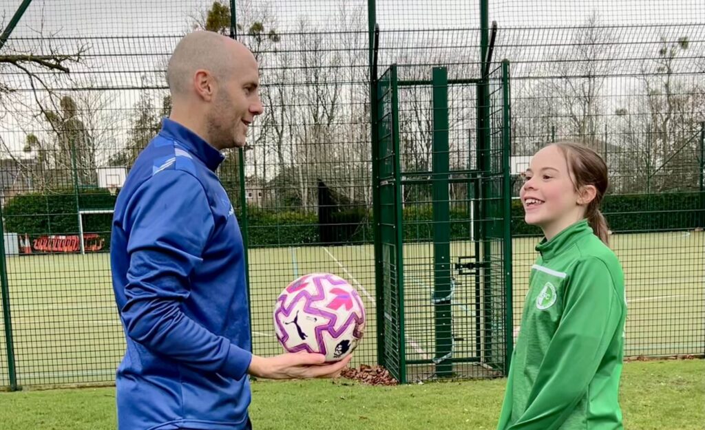Jordan wearing a blue top and blue leggings and holding a ball, speaking to a primary school age girl wearing a green top and blue legging