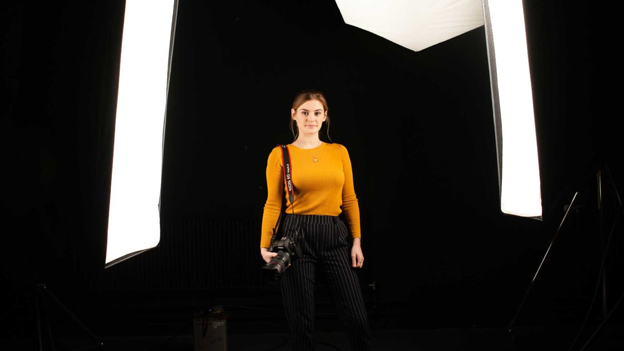 A photographer holding a DSLR camera stands between large lights in a photography studio.