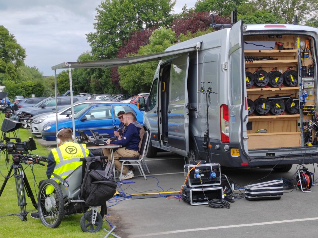 a silver van containing TV broadcasting equipment with two people sat in front of it at a table holding microphones